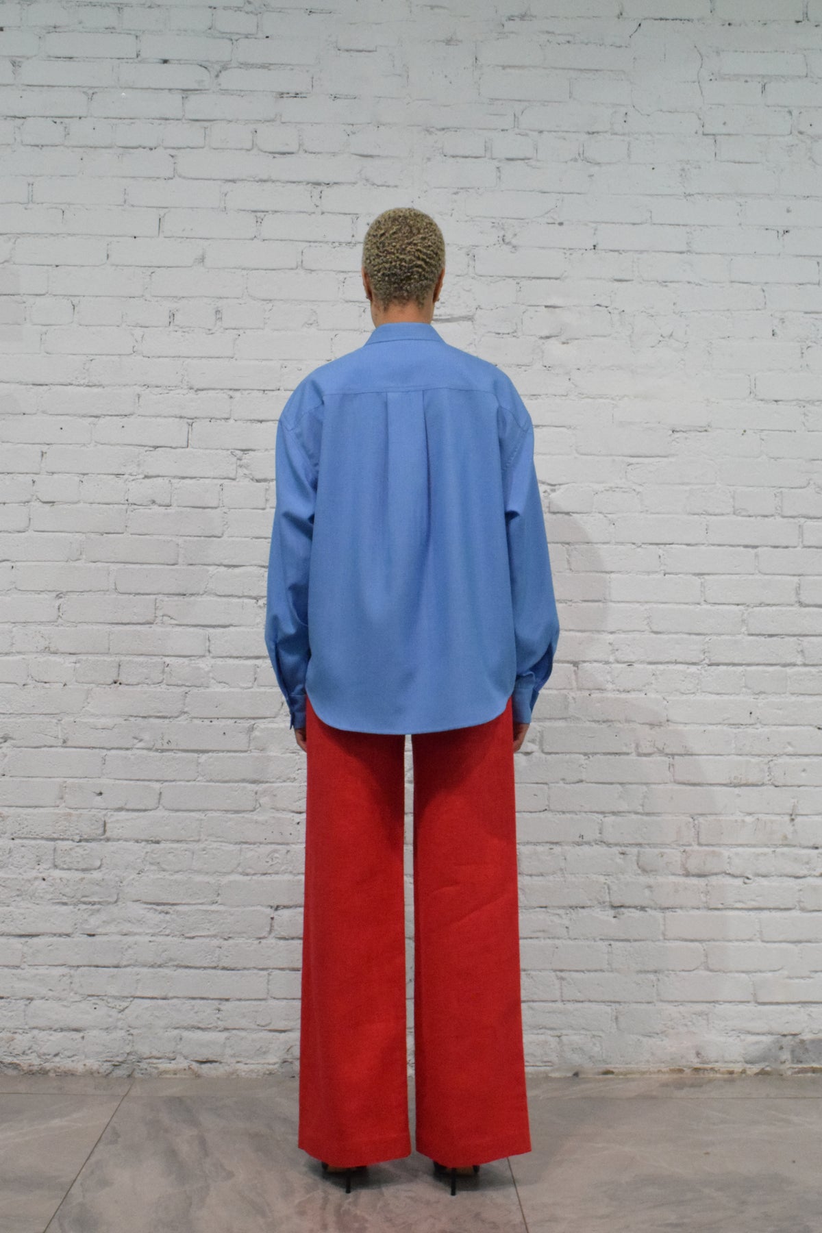 Person wearing a blue shirt and red pants standing against a white brick wall.