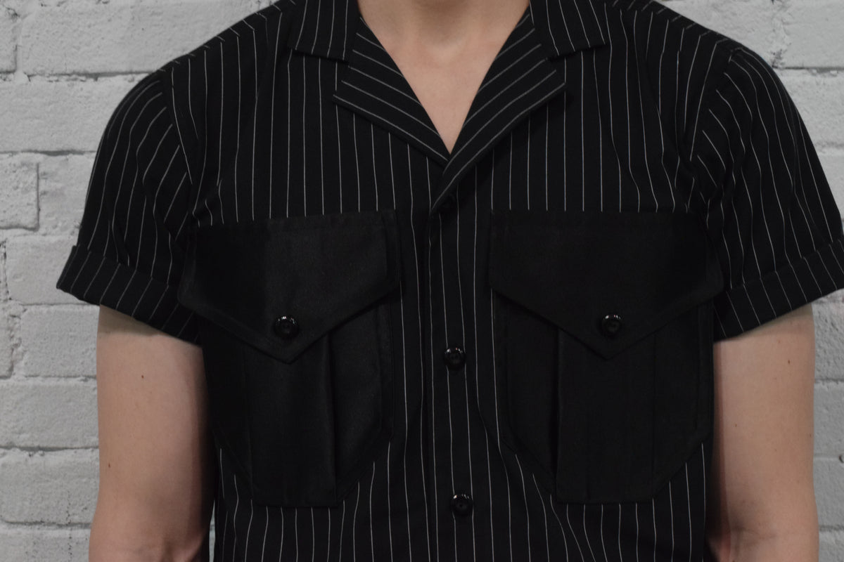 Black pinstripe shirt with short sleeves and button details against a gray brick wall.