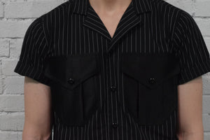 Black pinstripe shirt with short sleeves and button details against a gray brick wall.
